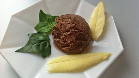 Ein Schokoladeneis in einer Schale, umgeben von frischen Blättern und gelben Fruchtstücken auf einem stilvollen Teller.