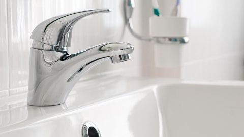 Ein glänzender Wasserhahn fließt über ein weißes Waschbecken in einem hellen Badezimmer mit Zahnbürsten im Hintergrund.