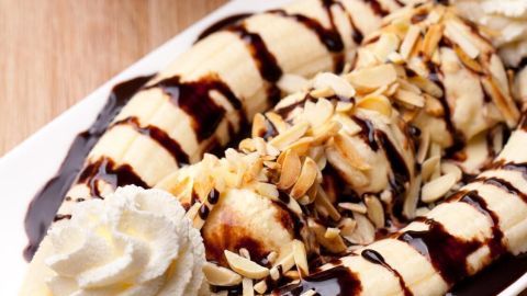 Bananensplit mit Schokoladensauce, Sahne und Mandeln auf weißem Teller, umgeben von frischen Bananen auf einem Holztisch.