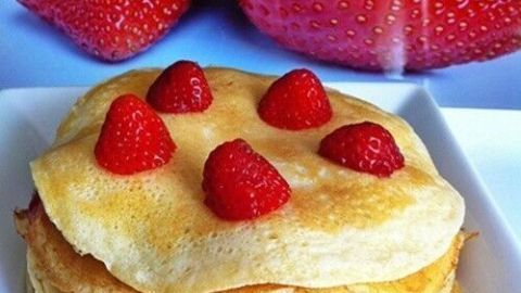 Gestapelte, goldbraune Pfannkuchen sind mit frischen Erdbeeren bedeckt, im Hintergrund sind große Erdbeeren zu sehen.