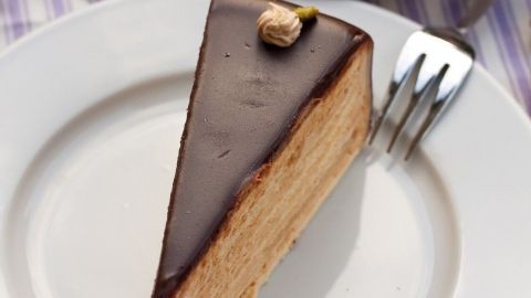Ein Stück Nougat-Sahne-Torte mit Knusperboden liegt auf einem weißen Teller, während eine Tasse Kaffee daneben steht.