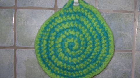 Ein gefilzter Topflappen in spiralförmigem Design hängt an einem Haken, umgeben von fliesenartigen Wänden.