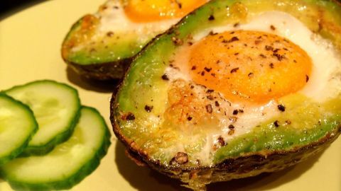 Zwei halbierte Avocados mit einem gebackenen Ei in der Mitte liegen auf einem gelben Teller, umgeben von Gurkenscheiben.