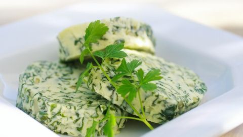 Grüne Bärlauchbutter liegt auf einem weißen Teller, dekoriert mit frischer Petersilie in einem hellen Küchenumfeld.