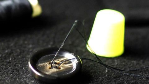 Ein schwarzer Knopf liegt auf dunklem Untergrund. Eine Nadel mit schwarzem Faden ist eingestochen. Ein gelber Fingerhut steht daneben.