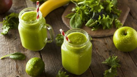Zwei Gläser gesunder grüner Smoothies stehen auf einem Holztisch, umgeben von frischen Früchten und Blättern.