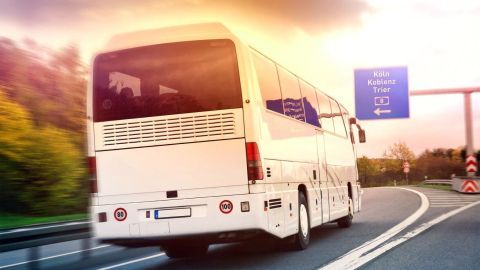 Ein Reisebus fährt schnell auf einer Autobahn, umgeben von grünen Bäumen und unter einem bunten Himmel.