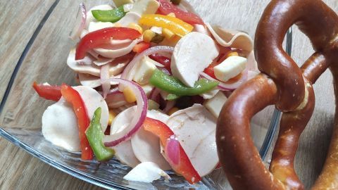 Ein bunter Wurstsalat wird in einer Glasschüssel präsentiert, umgeben von einer Brezel auf einem Holztisch.