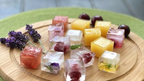 Bunte Eiswürfel aus Fruchtsaftresten liegen auf einem Holzbrett, umgeben von Lavendel und frischen Früchten.