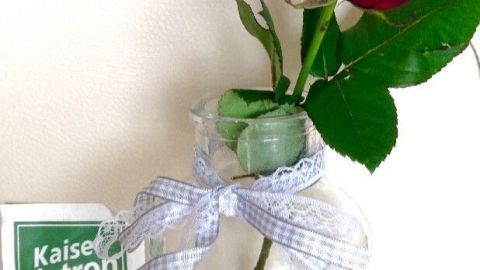 Eine rote Rose steht in einer dekorativen Vase mit einem Band, neben einem Päckchen Kaiser Natron für längere Frische.