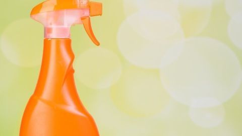 Eine orangefarbene Sprühflasche steht neben einem pink-blauen Schwamm auf einer gelben Oberfläche mit unscharfem Hintergrund.