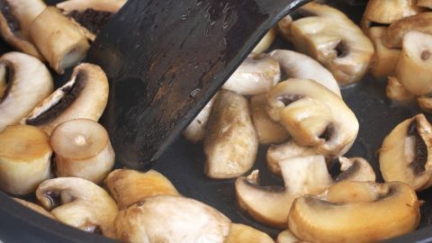 Champignons werden in einer Pfanne gebraten, während ein Spatel die Pilze vorsichtig bewegt. Eine Küchenumgebung ist sichtbar.