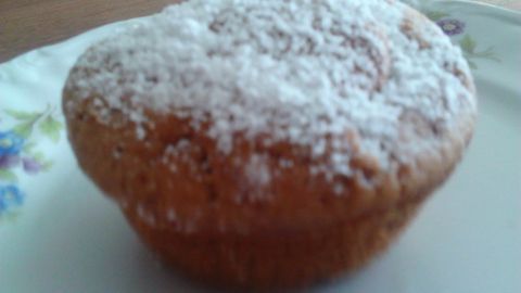 Ein süßer Muffin mit Puderzucker bestreut steht auf einem Blumen-musternden Teller im hellen, einladenden Raum.