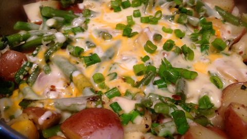 Eine bunte Kartoffelpfanne mit grünen Bohnen und geschmolzenem Käse wird in einer Pfanne serviert, umgeben von frischen Kräutern.