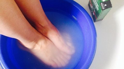 Zwei Füße sind in einem blauen Behälter mit Wasser eingetaucht, neben dem steht eine Packung Kaiser Natron für das Fußbad.