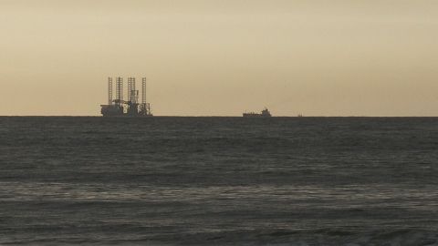 Eine Bohrinsel steht stabil im Wasser, während ein kleines Schiff vorbeifährt, unter einem sanften Sonnenuntergang.