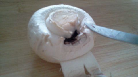 Ein Champignon wird mit einem Messer schnell und trocken gereinigt. Die Umgebung ist eine Holzoberfläche.