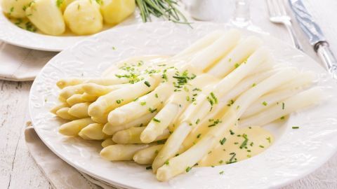 Weißer Spargel liegt auf einem Teller, in Buttersoße serviert, umgeben von Kartoffeln auf einem Tisch, alles schön angerichtet.