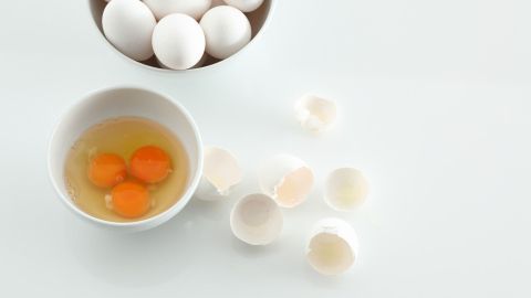 Eine Schüssel mit drei rohen Eiern neben zerkleinerten Eierschalen und einer weiteren Schüssel voller Eier auf einem weißen Tisch.