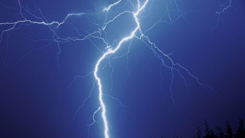 Ein heller Blitz zuckt durch den dunklen Himmel während eines Gewitters, umgeben von düsteren Wolken und silhouettenhaften Bäumen.