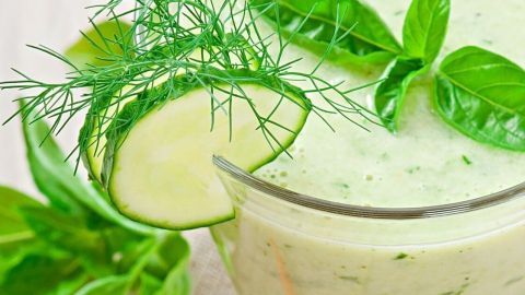 Ein Gurkensmoothie steht auf einem Tisch, garniert mit Basilikum und Dill, daneben liegen Chilis und ein Brotstück.