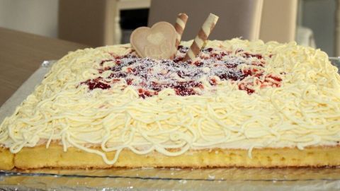 Ein Spaghetti Kuchen liegt auf einem Tisch. Die Optik wird durch weiße Creme und rote Soße ergänzt, verziert mit Waffelröllchen und Keksen.