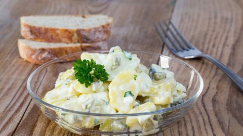 Ein bunter Kartoffelsalat mit selbst gemachter Mayonnaise liegt in einer Glasschüssel auf einem Holztisch, daneben Brot und eine Gabel.