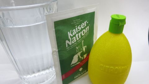 Selbst gemachte Brause mit Natron und Zitronensäure: Lecker im Sommer, da Fertigprodukte oft sehr süß sind. Ein gelbes Flaschenobjekt und ein Päckchen Brausepulver stehen neben einem Wasserglas und deuten auf das Mixen eines Erfrischungsgetränks hin.