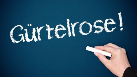 Eine Hand schreibt "Gürtelrose!" mit einem weißen Kreidestift auf eine schwarze Tafel, um das Thema visuell zu betonen.
