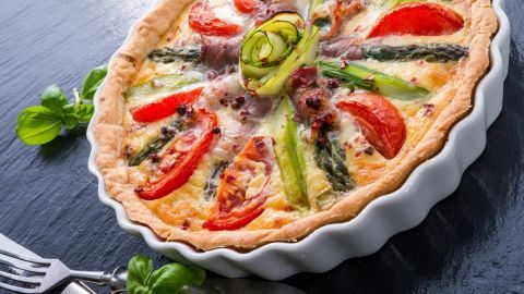 Eine frisch gebackene Quiche mit grünem Spargel und Tomaten liegt dekorativ in einer weißen Tarteform auf einem dunklen Tisch.