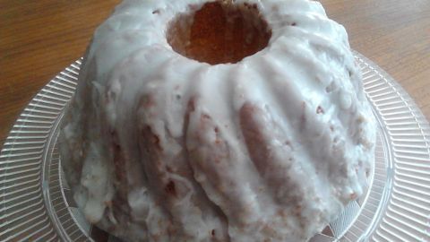 Ein köstlicher Zitronenkuchen mit glasiertem Überzug steht auf einem Durchsichtigen, runden Teller vor einem Holzuntergrund.
