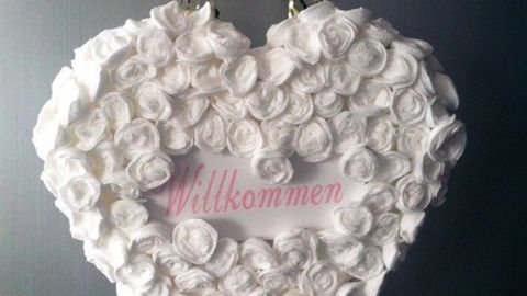 Ein herzförmiger Türschmuck aus weißen Rosenblüten hängt an einer Kette, mit dem Schriftzug "Willkommen" in der Mitte.