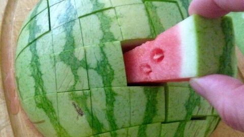 Eine auf einem Holzbrett liegende Wassermelone wird in Stiftform geschnitten, während ein Stück herausgenommen wird.