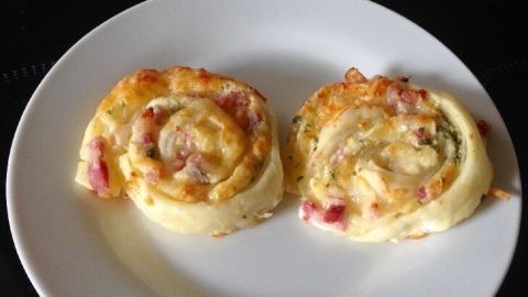 Zwei Blätterteigschnecken mit Schmand und Bacon liegen auf einem weißen Teller und sehen goldbraun und knusprig aus.