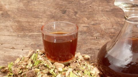 Ein Glas mit einer dunklen Flüssigkeit steht vor einer Menge getrockneter Kräuter, die für Schwedenkräuter gegen Pickel verwendet werden.