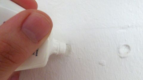 Kleine Bohrlöcher werden mit weißer Zahnpasta wieder unsichtbar! Eine Hand hält eine Tube Zahnpasta, während sie versucht, Bohrlöcher in einer weißen Wand zu füllen.