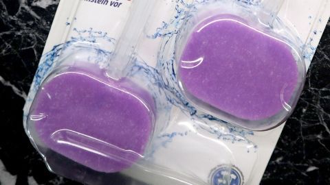Ein Klostein im Motorraum kann gegen Marderprobleme helfen. Zwei lila Klosteine sind in einer transparenten Verpackung abgebildet, umgeben von Wasserspritzern auf schwarzem Marmor.