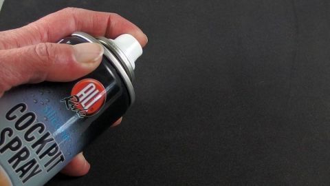 Eine Hand hält eine Spraydose mit Cockpitspray, die auf eine dunkle Oberfläche gerichtet ist, um eine Maus zu reinigen.