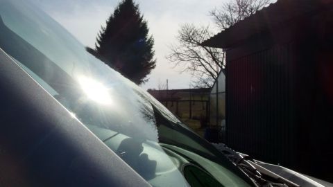 Ein Auto mit einer gewellten Windschutzscheibe steht hell erleuchtet im Freien, umgeben von Bäumen und einem Schuppen.