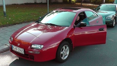 Ein rotes Auto hält an, die Fahrertür ist geöffnet, während eine Person im Fahrersitz sitzt und aussteigt.
