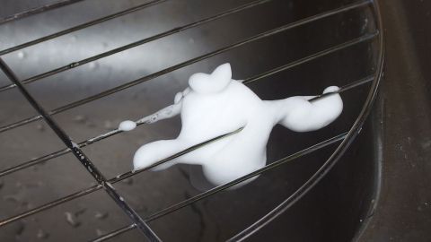 Ein Grillrost liegt auf einem Tisch, w&auml;hrend wei&szlig;e Schaumreinigungsmittel darauf aufgetragen werden, um ihn zu s&auml;ubern.