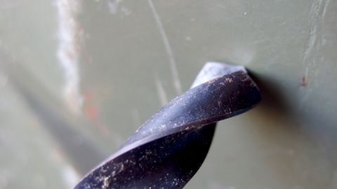 Mit einem Stückchen Holz kann man sich das Bohren in Blech sehr vereinfachen. Ein Bohrer dreht sich, während er in ein Blech eindringt, um präzise Löcher zu erzeugen. Der Kontext zeigt eine metallische Oberfläche.