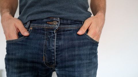 Eine Person steht mit beiden Händen in den Taschen ihrer Jeans, vor einem neutralen, hellen Hintergrund.
