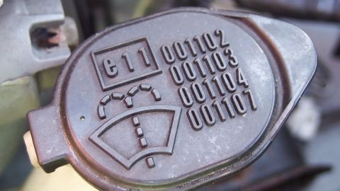 Der Einsatz von Scheibenwaschzusatzmittel gegen Schmutz, Fettschlieren und Fliegenresten während der Autofahrt, bringt meist wenig. Hier eine preiswerte Alternative, die tatsächlich Erfolg bringt. Ein schwarzes Behälterdeckel zeigt Symbole und Zahlen für Scheibenwaschmittel, montiert im Motorraum eines Fahrzeugs.