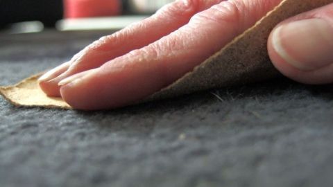 Hand reibt mit Sandpapier über Teppich, um Tierhaare zu entfernen. Nahaufnahme zeigt Detailarbeit in häuslicher Umgebung.