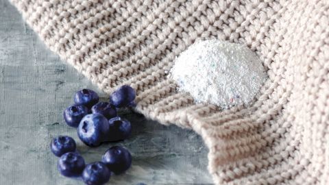 Ein gestricktes Kleidungsstück mit einem Blaubeerfleck und daneben liegen frische Blaubeeren auf einer Oberfläche.