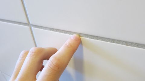 Eine Hand mit schön manikürten Fingernägeln berührt eine glatte, weiße Fliese an der Wand in einem hellen Raum.