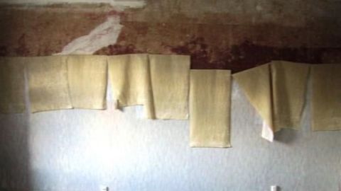 Tapeten hängen halb abgelöst von einer Wand, der freigelegte Putz ist sichtbar, während der Boden den Raum kontextualisiert.