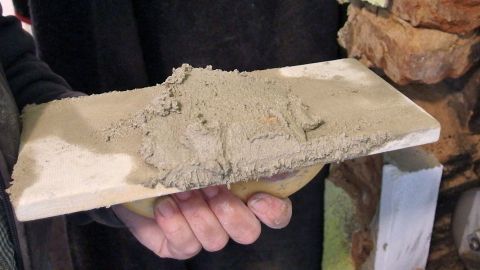 Eine Hand hält eine Kelle mit frischem Mauermörtel, während sich im Hintergrund eine Steinmauer befindet.