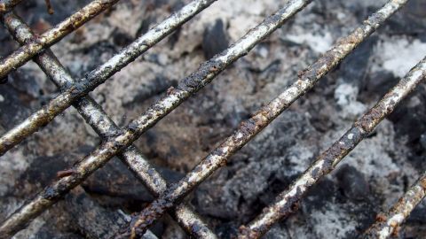 Ein stark verschmutzter Grillrost liegt &uuml;ber verkohltem Grillgut, umgeben von Asche und R&uuml;ckst&auml;nden, die eine Reinigung ben&ouml;tigen.